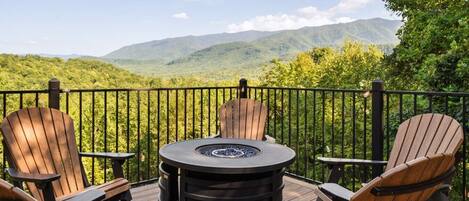 Eine Terrasse mit Adirondack-Stühlen und einem runden Tisch mit Feuerstelle, mit Blick auf eine gebirgige Landschaft.