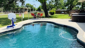 Une piscine dotée d’un panier de basketball, entourée d’une zone délimitée par une clôture et comprenant des sièges extérieurs.