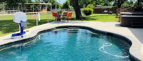 Una piscina con aro de baloncesto, rodeada por un área cercada con mobiliario exterior.