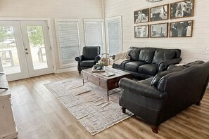 A living room with a dark leather sofa, a wooden coffee table, and a patterned rug. There are two chairs, a fireplace, and a set of framed pictures on the wall.