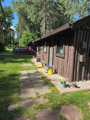 A wooden cabin with a paved walkway leading to it, surrounded by greenery and a parked car.