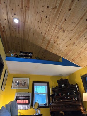 A room with a wooden ceiling, a window with a view, a radiator, a mirror, and a piano.