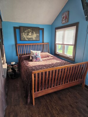 A bedroom with a wooden bed, a small table, a window with blinds, and a wall-mounted picture. - Cozy 1-bedroom cabin in amazing White Lake with AC on ALL SPORTS SUGDEN LAKE (White Lake)