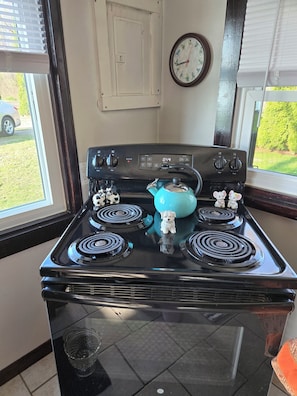 A kitchen with a stove, a clock, and a window with a view of the outside.
