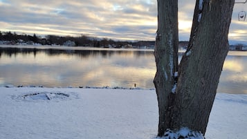 A snow-covered outdoor seating area with a bench and table, overlooking a calm lake with reflections of trees and houses in the distance.