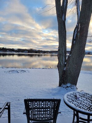 A snow-covered outdoor seating area with a bench and table, overlooking a calm lake with reflections of trees and houses in the distance.