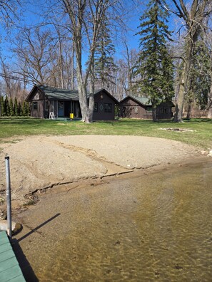 A cabin with a deck, a sandy area, and a clear water body.