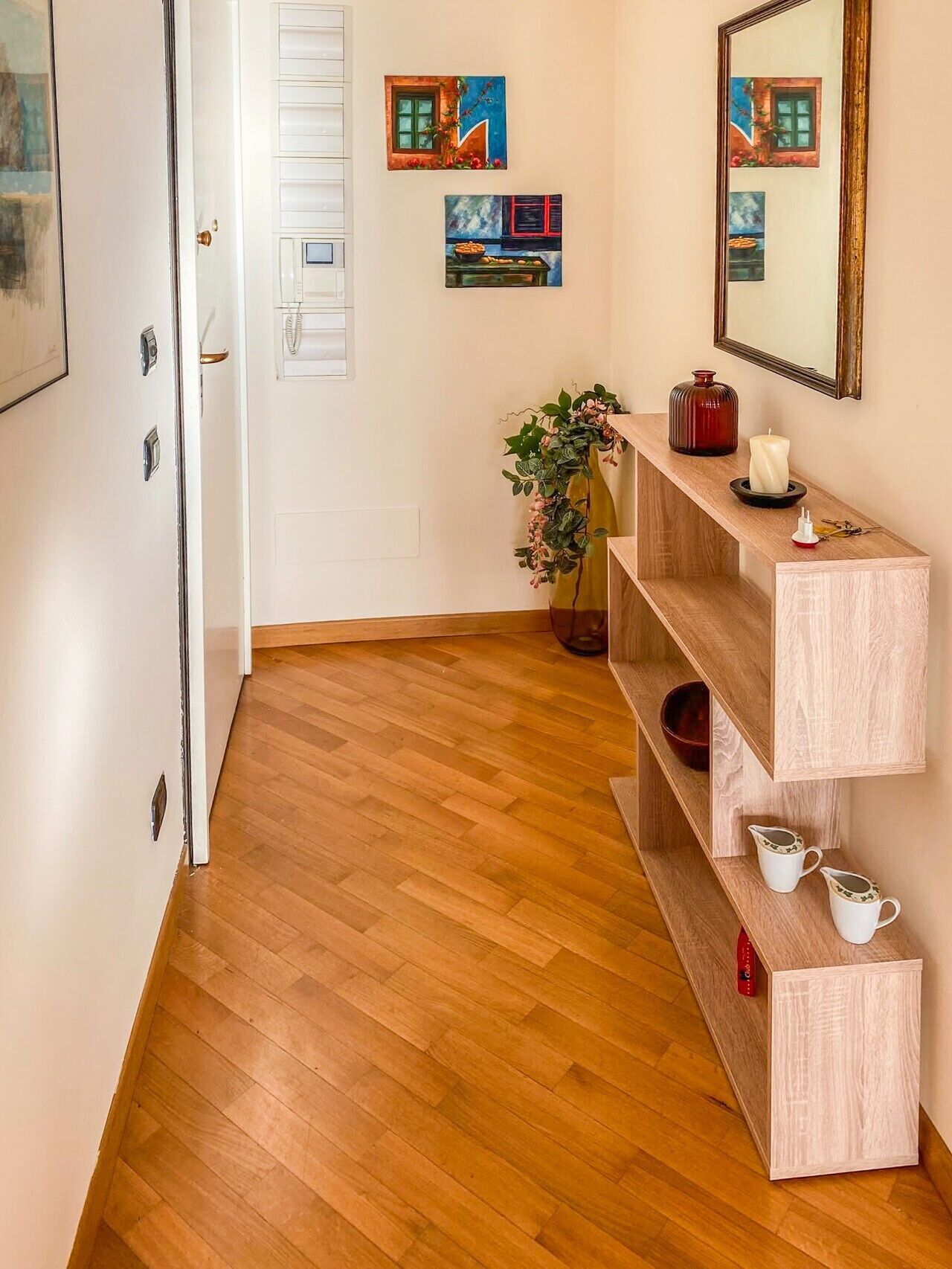 Un couloir doté d’un plancher en bois, d’une porte blanche, d’une console en bois ornée d’objets décoratifs et d’œuvres d’art accrochées au mur.