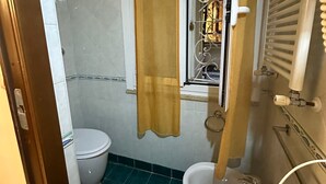 A small bathroom with a toilet, a bidet, a shower area, and a window with a curtain.