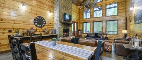 A log cabin interior with a dining area, fireplace, leather seating, and large windows.