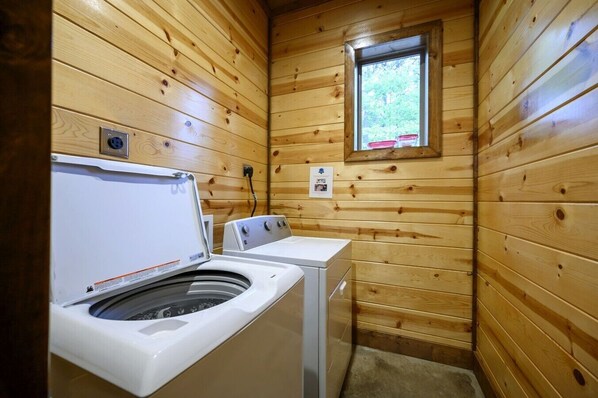 A compact laundry room with wooden walls, a washing machine, a dryer, and a window providing natural light.