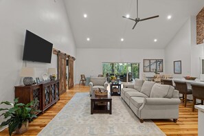 A spacious living room with a large flat-screen TV, a ceiling fan, and a dining area with a view of the outdoors. - Lakefront MASTERS WEEK Rental (McCormick)