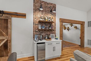 A kitchen with a brick wall, wooden shelves with bottles, a white cabinet, and a stainless steel refrigerator. - Lakefront MASTERS WEEK Rental (McCormick)