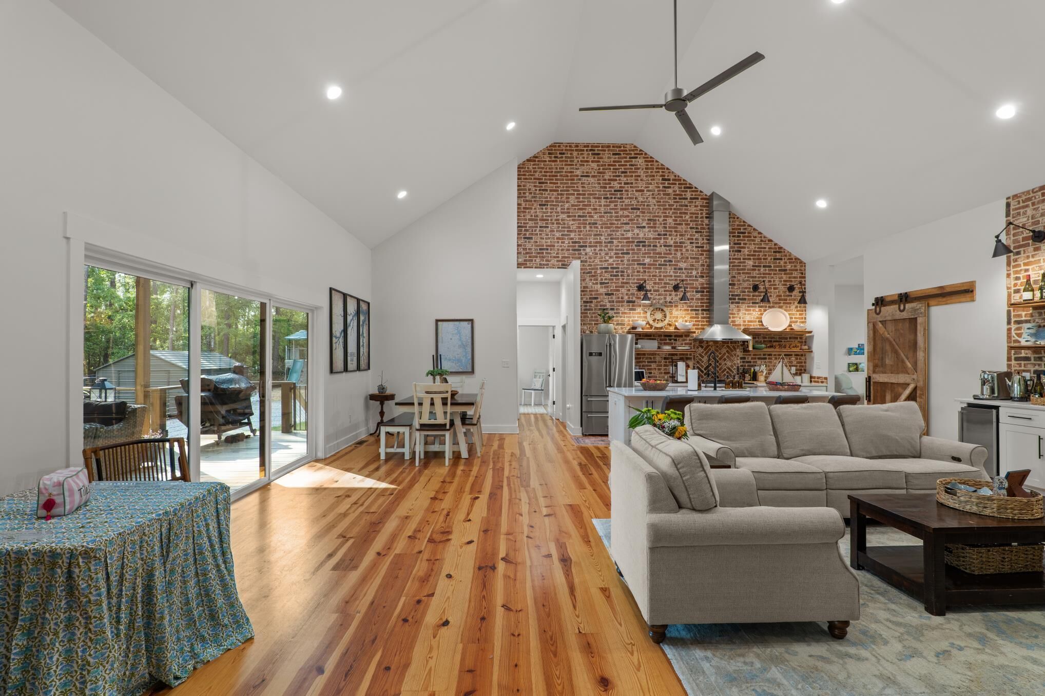 A spacious living area with a wooden floor, a large sofa, a dining table, and a kitchen with a brick wall.
