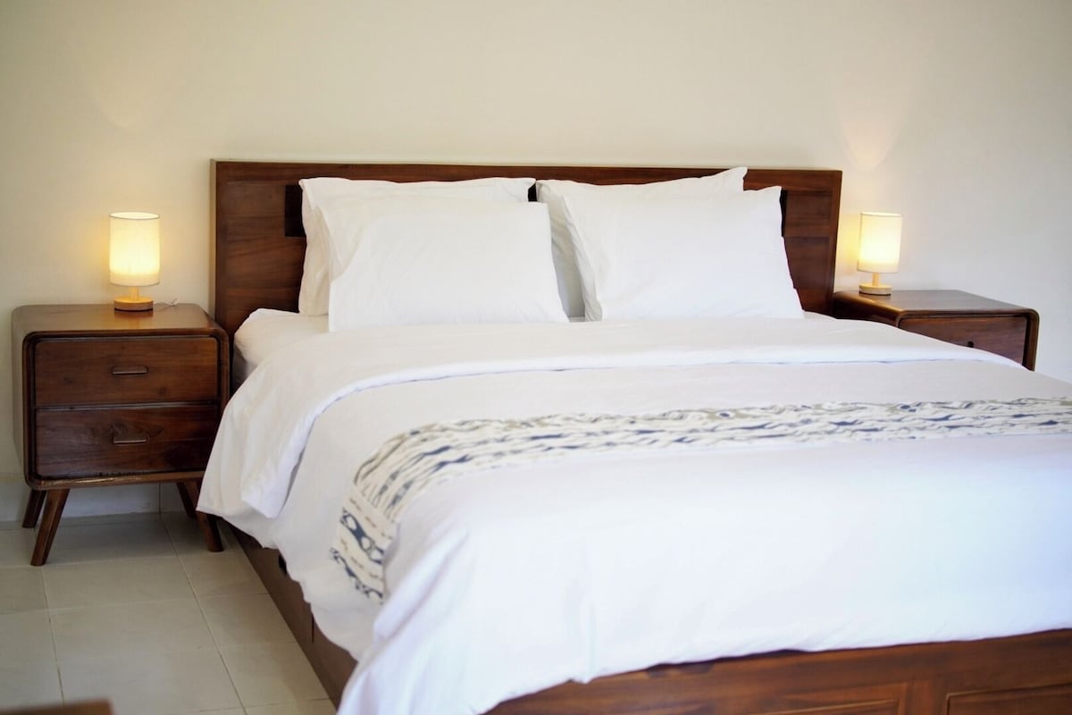 A neatly made bed with white linens and a patterned throw, flanked by two bedside tables with lamps, in a room with a plain wall and wooden headboard.