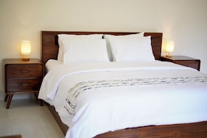 A neatly made bed with white linens and a patterned throw, flanked by two bedside tables with lamps, in a room with a plain wall and wooden headboard. - Dhanda Homestay (Ubud)