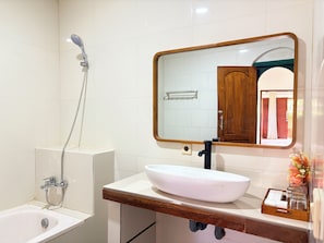 A bathroom with a white bathtub, a white sink with a wooden countertop, a mirror with a wooden frame, and a shower with a handheld showerhead. - Dhanda Homestay (Ubud)
