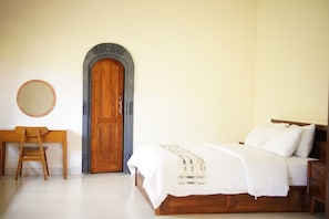 A bedroom with a wooden bed, a desk with a chair, a round mirror, and a wooden door with an arch. - Dhanda Homestay (Ubud)