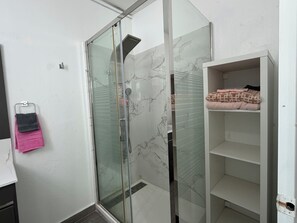 Une salle de bain avec une douche à parois de verre, un mur en marbre blanc et une étagère où sont pliées des serviettes.