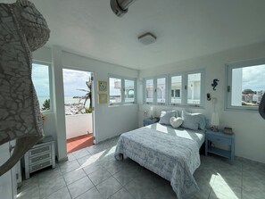 Une chambre à coucher avec un lit, des tables de chevet, une commode et une vue sur la mer grâce à de grandes fenêtres.