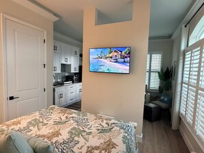 A bedroom with a bed, a TV on the wall, a kitchen in the background, and a window with blinds. - Serene & Luxurious Beach Studio, Heated Pool, Garage & Steps to Sand (St. Pete Beach)
