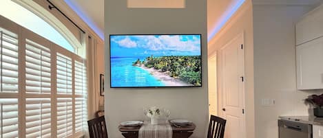 Un comedor con una mesa dispuesta para dos, un televisor de pantalla plana montado encima y una cocina contigua al área de comedor.