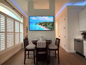 A dining area with a table set for two, a flat-screen TV mounted above it, and a kitchen adjacent to the dining space.