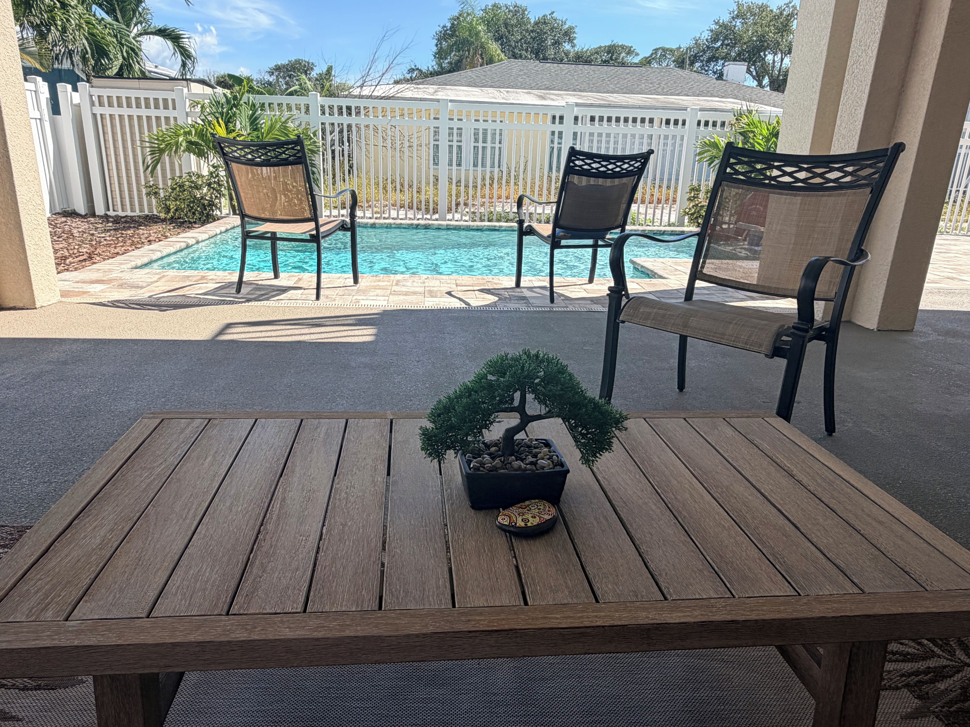 A wooden table with a bonsai tree centerpiece, surrounded by two chairs with mesh backs, a pool, and a white fence in the background.