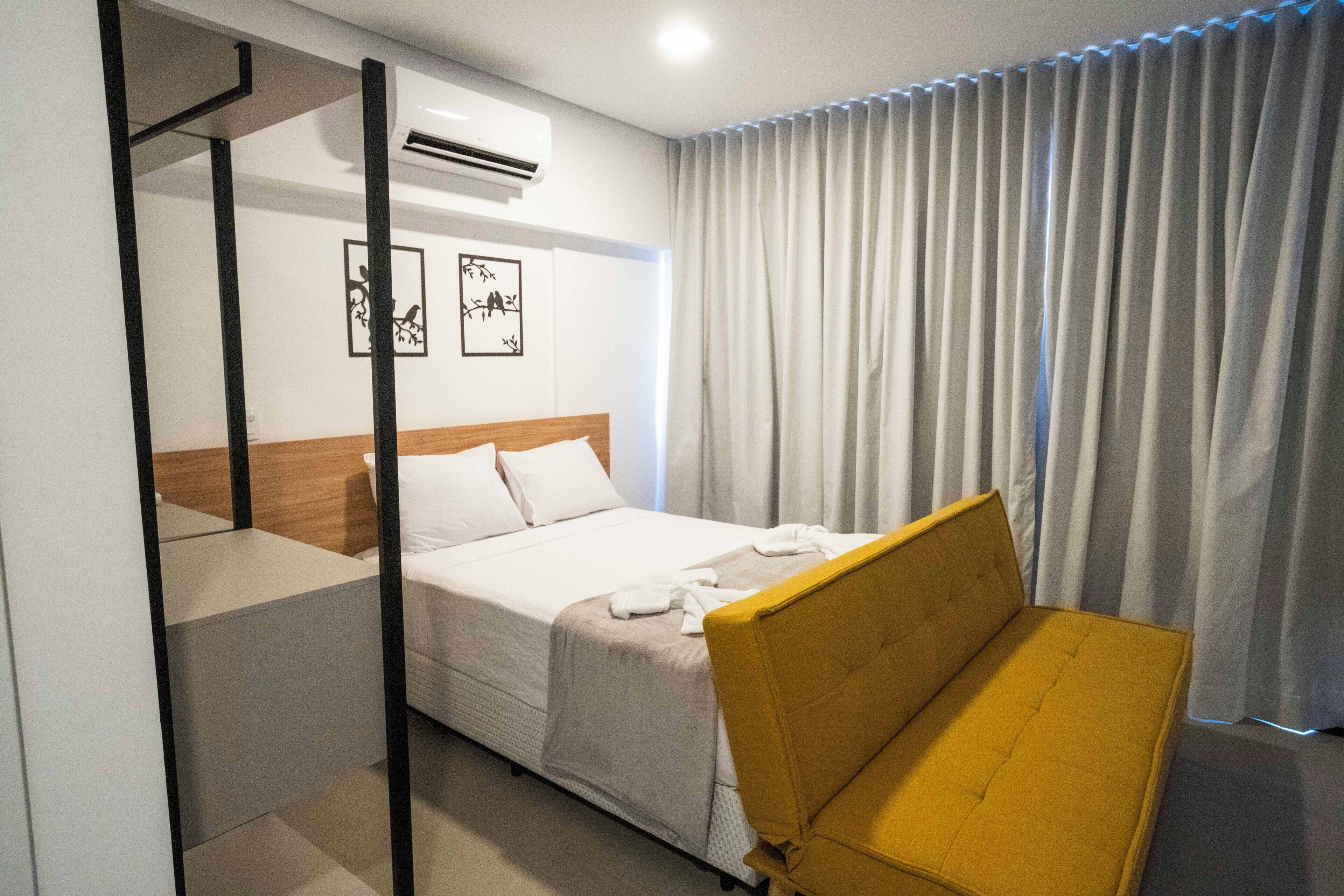 Une chambre d’hôtel avec un lit, une chaise jaune, un appareil de climatisation et deux œuvres d’art murales.