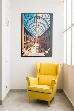 Une pièce avec un fauteuil jaune, un tableau encadré représentant un bâtiment historique et une fenêtre.