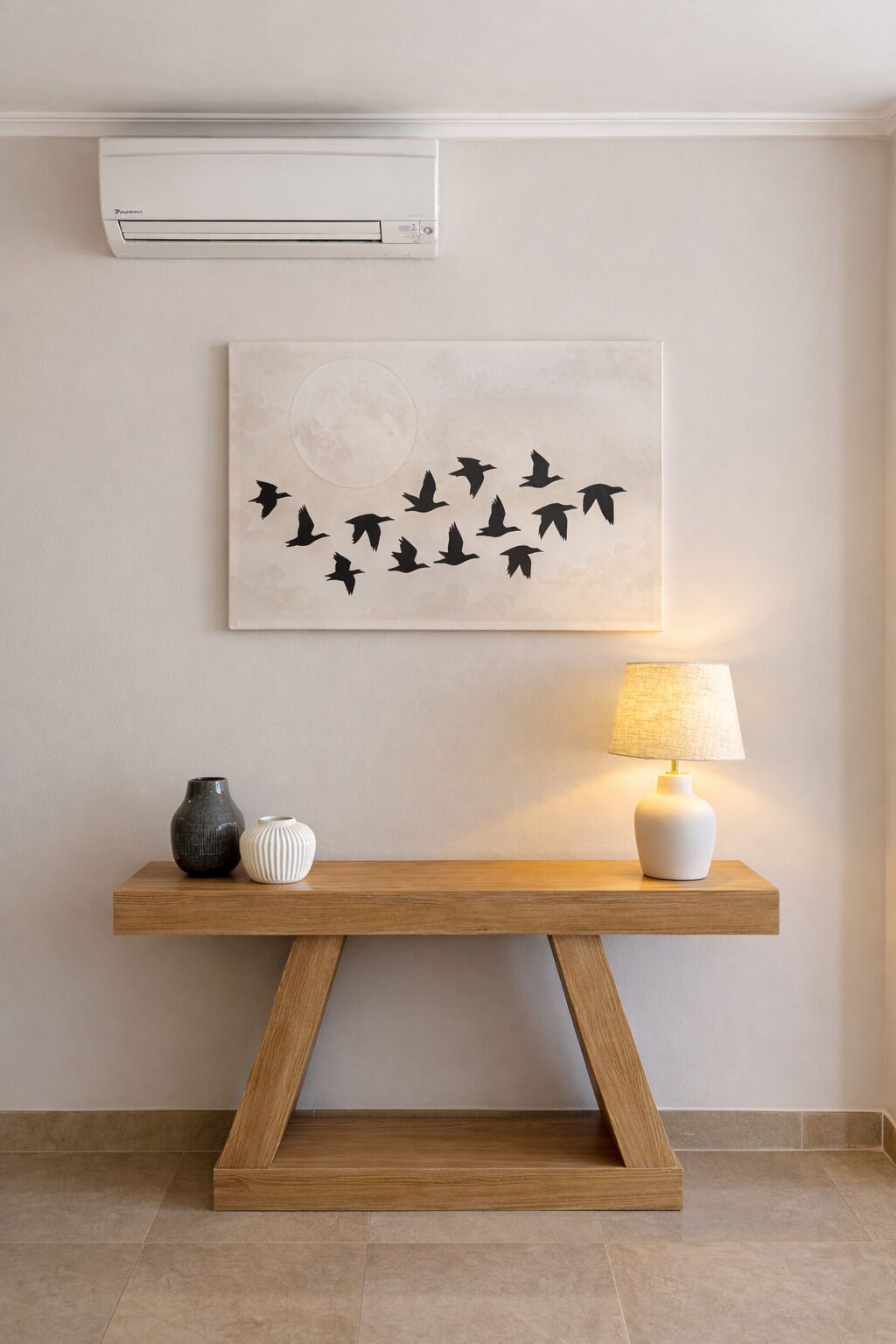 Una habitación con una consola de madera, un aire acondicionado fijado a la pared, una obra de arte enmarcada con pájaros, una lámpara y dos jarrones.