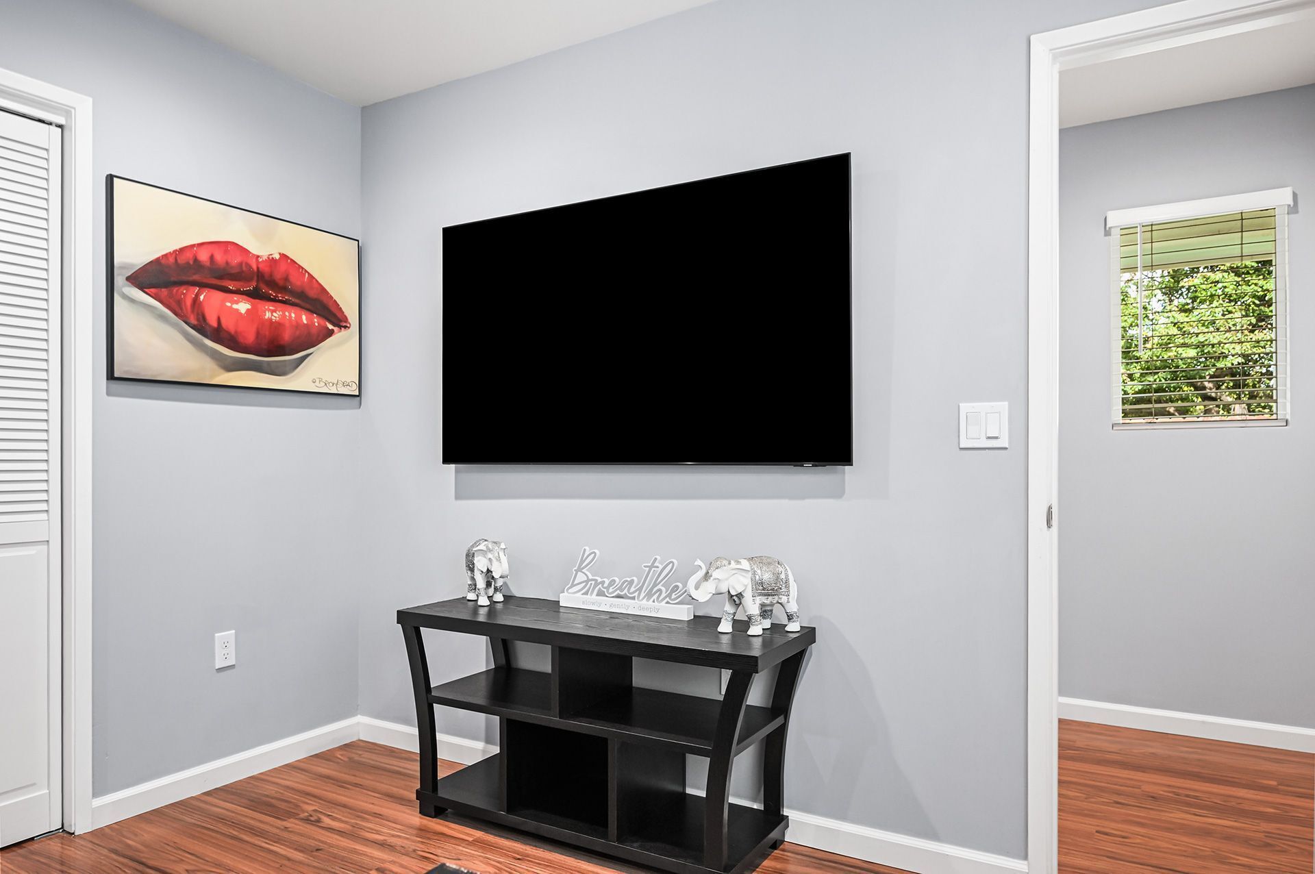 A room with a mounted flat-screen TV, a framed artwork of lips, a black console table with figurines, and a window with blinds.