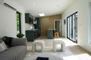 A modern living space with a sofa, ottomans, a kitchen area, and a sliding glass door leading to a balcony. - Modern Scandi Home on a Private Estate (Harbor Springs)