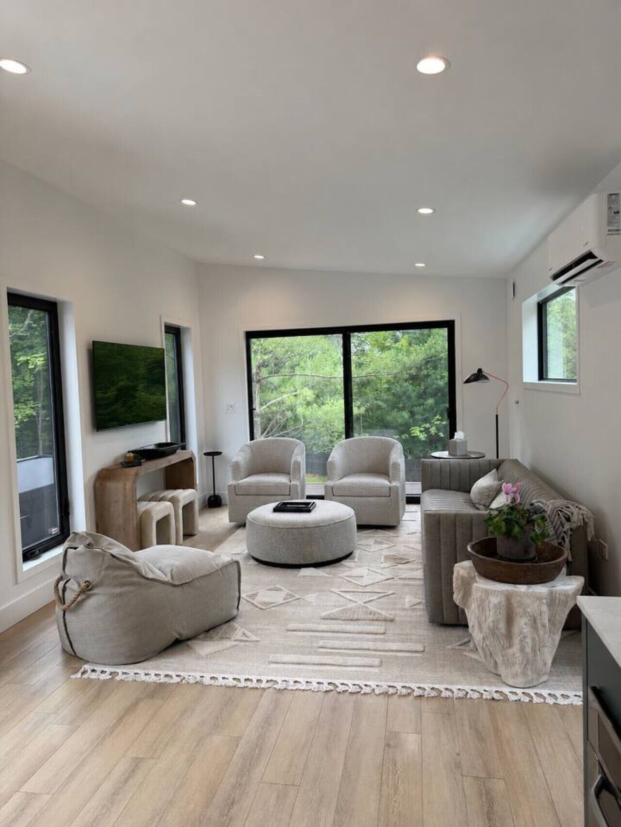 Una sala moderna con sofá, sillones, reposapiés y una mesa de centro. Amplias ventanas ofrecen luz natural y vistas al verdor.