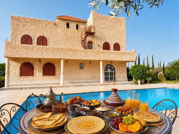 A poolside breakfast setup with a variety of foods and beverages, a traditional building with arched windows, and a clear blue sky.