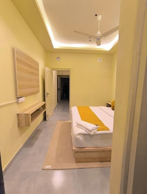 A hotel room with a bed, a ceiling fan, a wooden shelf, and a door leading to a hallway. - The Orchid Premium Studio By Verve (Greater Noida)