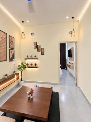 A modern dining area with a wooden table, cushioned chairs, pendant lighting, and wall-mounted shelves with decorative items.
