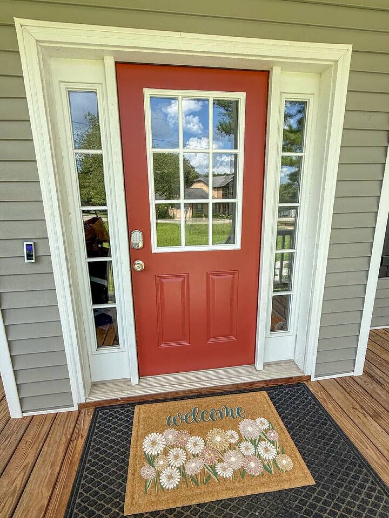 A red door with a white frame, a welcome mat with floral designs, and a small window on the door.
