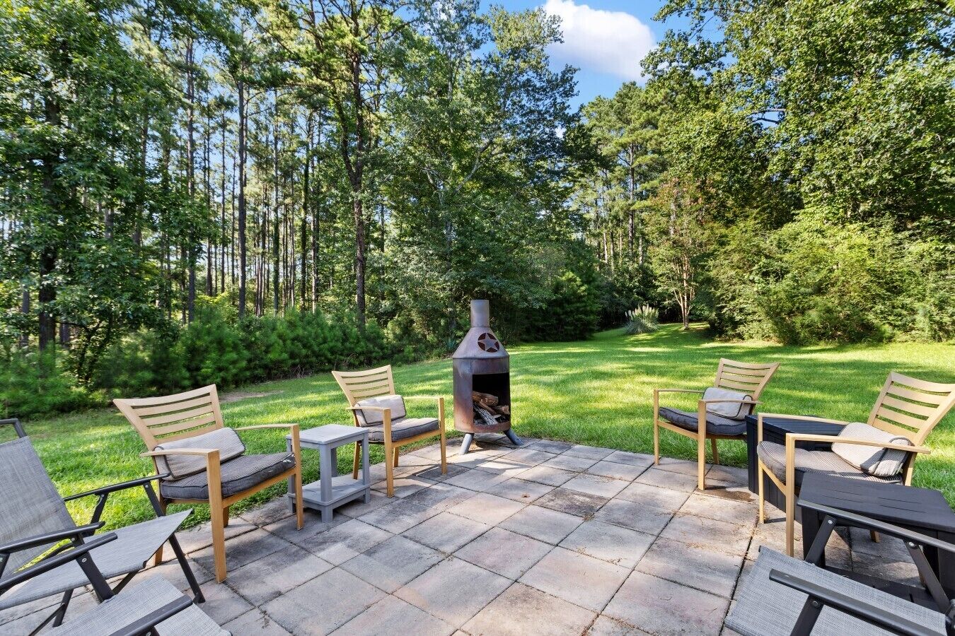 Une terrasse extérieure avec un sol en pierre, entourée d’une pelouse verdoyante et luxuriante et de grands arbres.