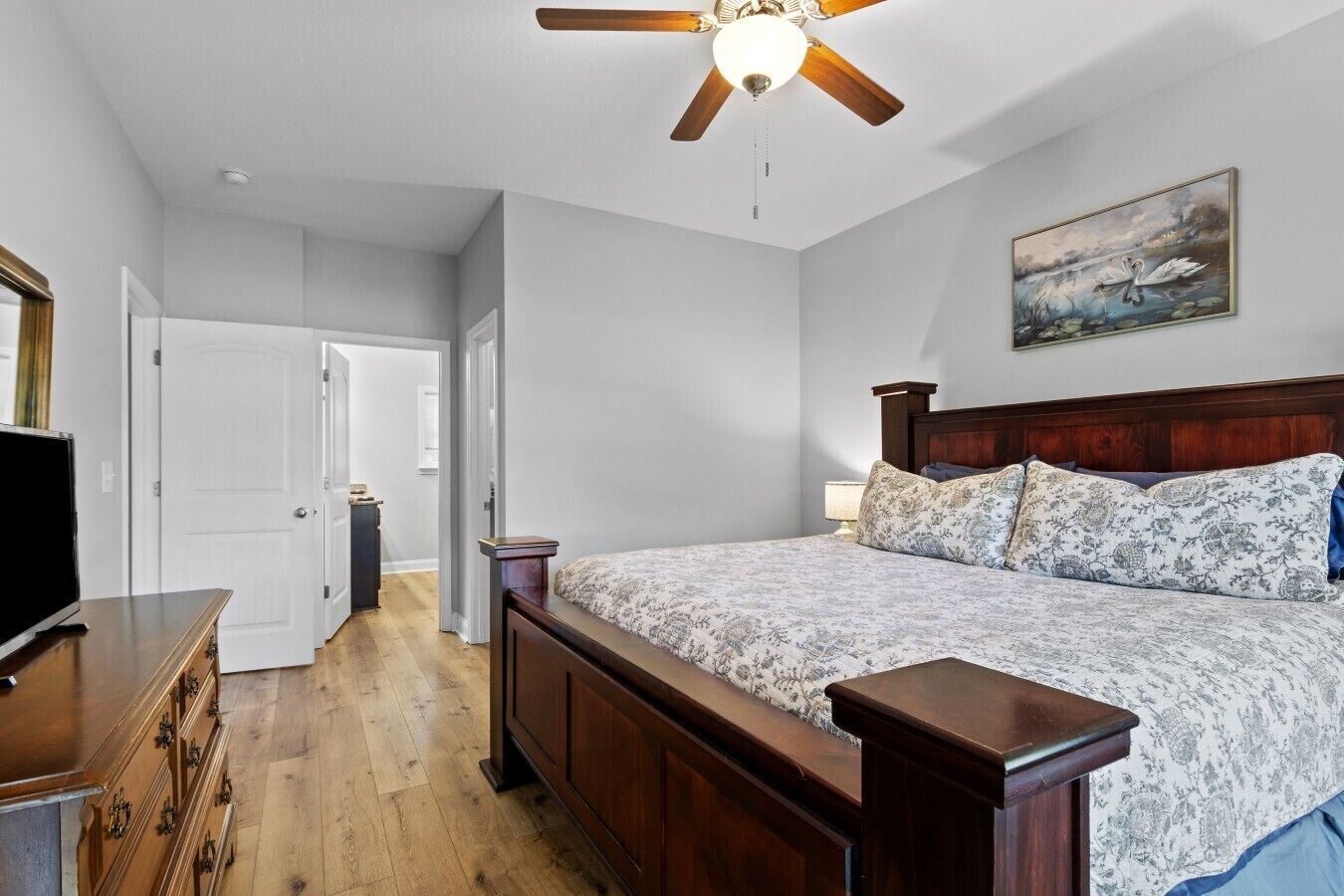 A bedroom with a wooden bed, a ceiling fan, a painting of swans, and a television on a dresser.