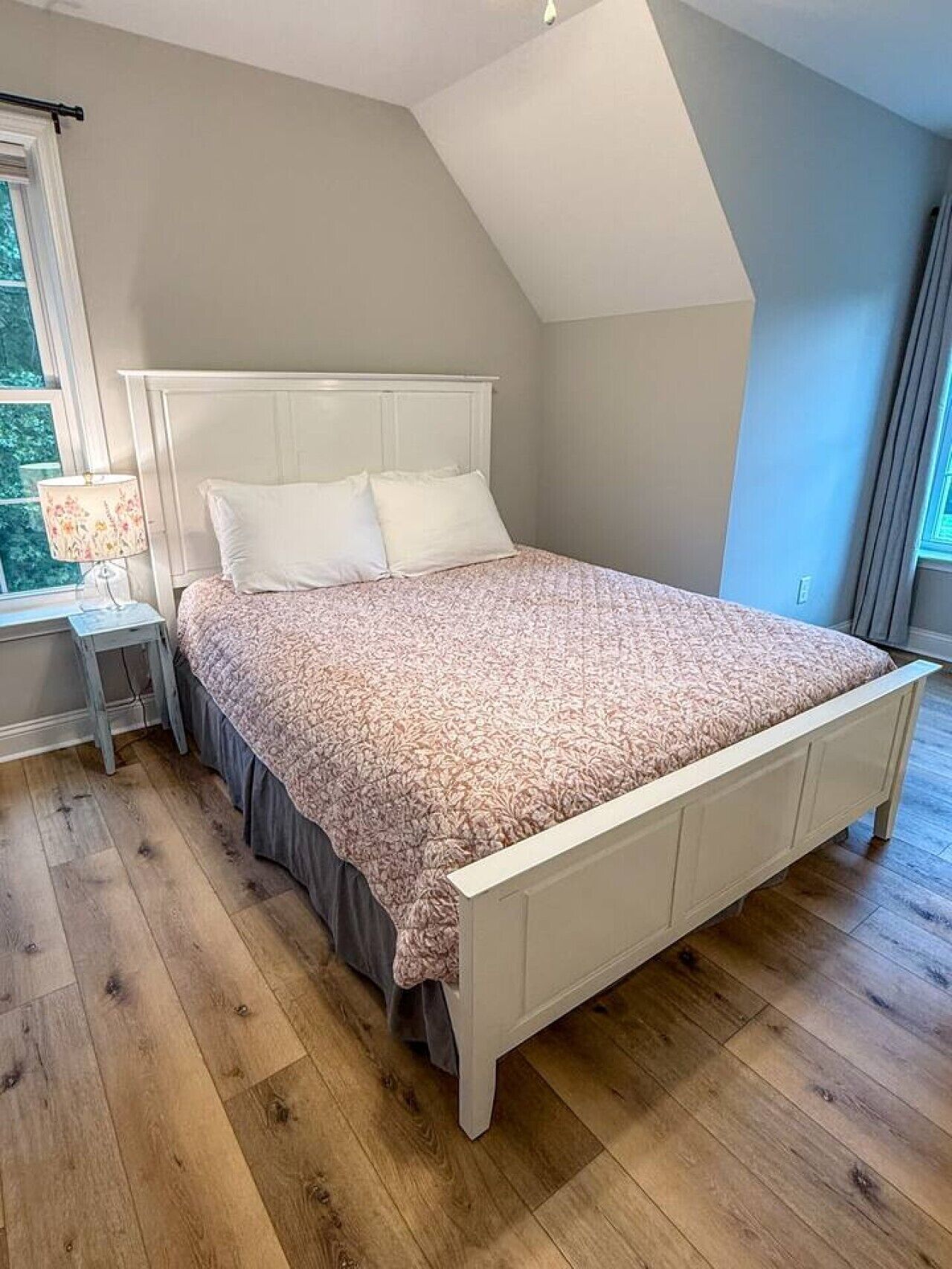 Ein Schlafzimmer mit einem weißen Bett, einem Nachttisch, einer Lampe und einem Fenster mit Vorhängen.
