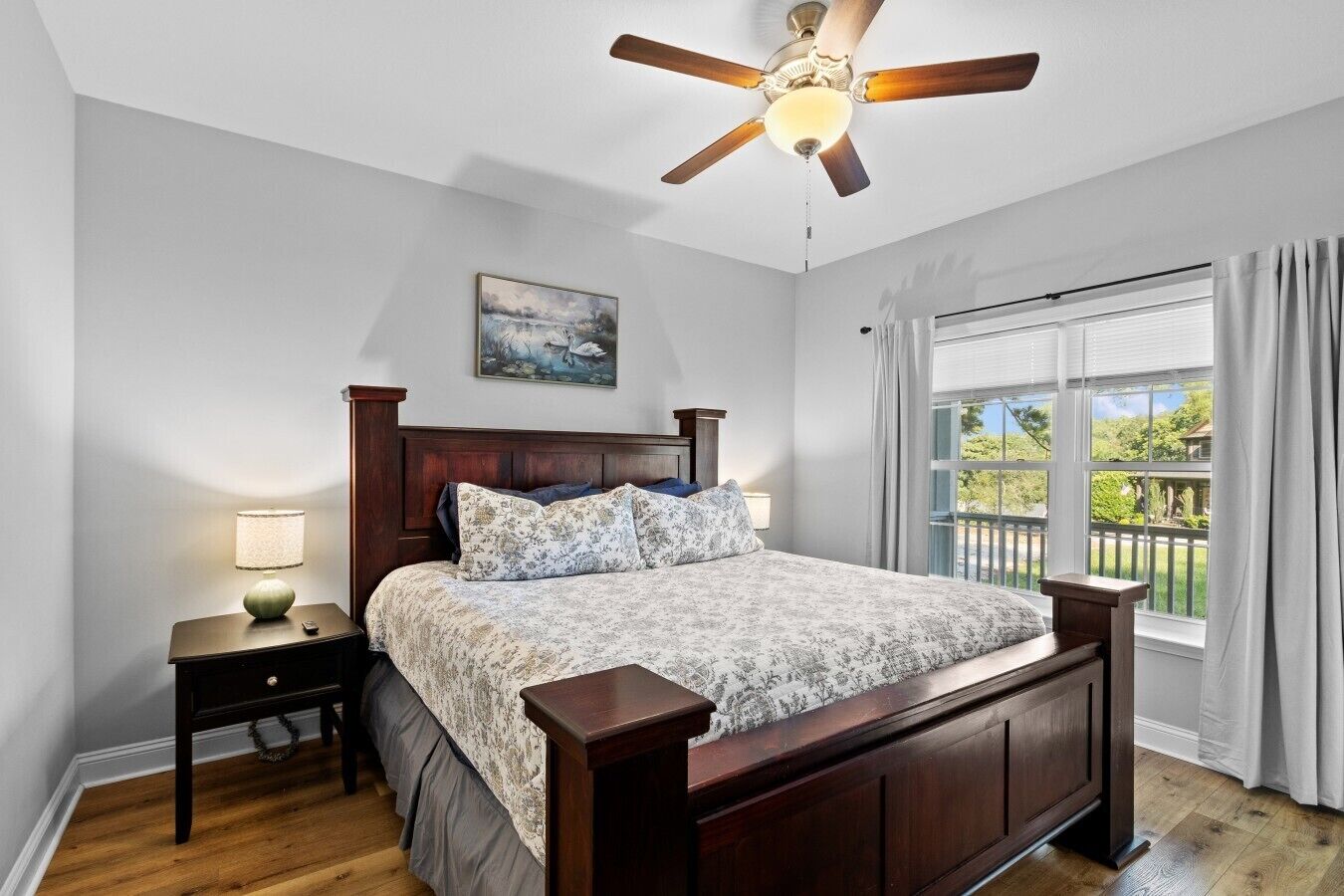 Une chambre avec un lit en bois, une table de chevet avec une lampe, un ventilateur de plafond, une fenêtre avec des rideaux et un tableau accroché au mur.