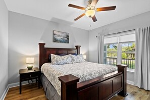 A bedroom with a wooden bed, a nightstand with a lamp, a ceiling fan, a window with curtains, and a painting on the wall. - Walk to Boat Ramp Lake Keowee w/ Parking + Firepit (Seneca)