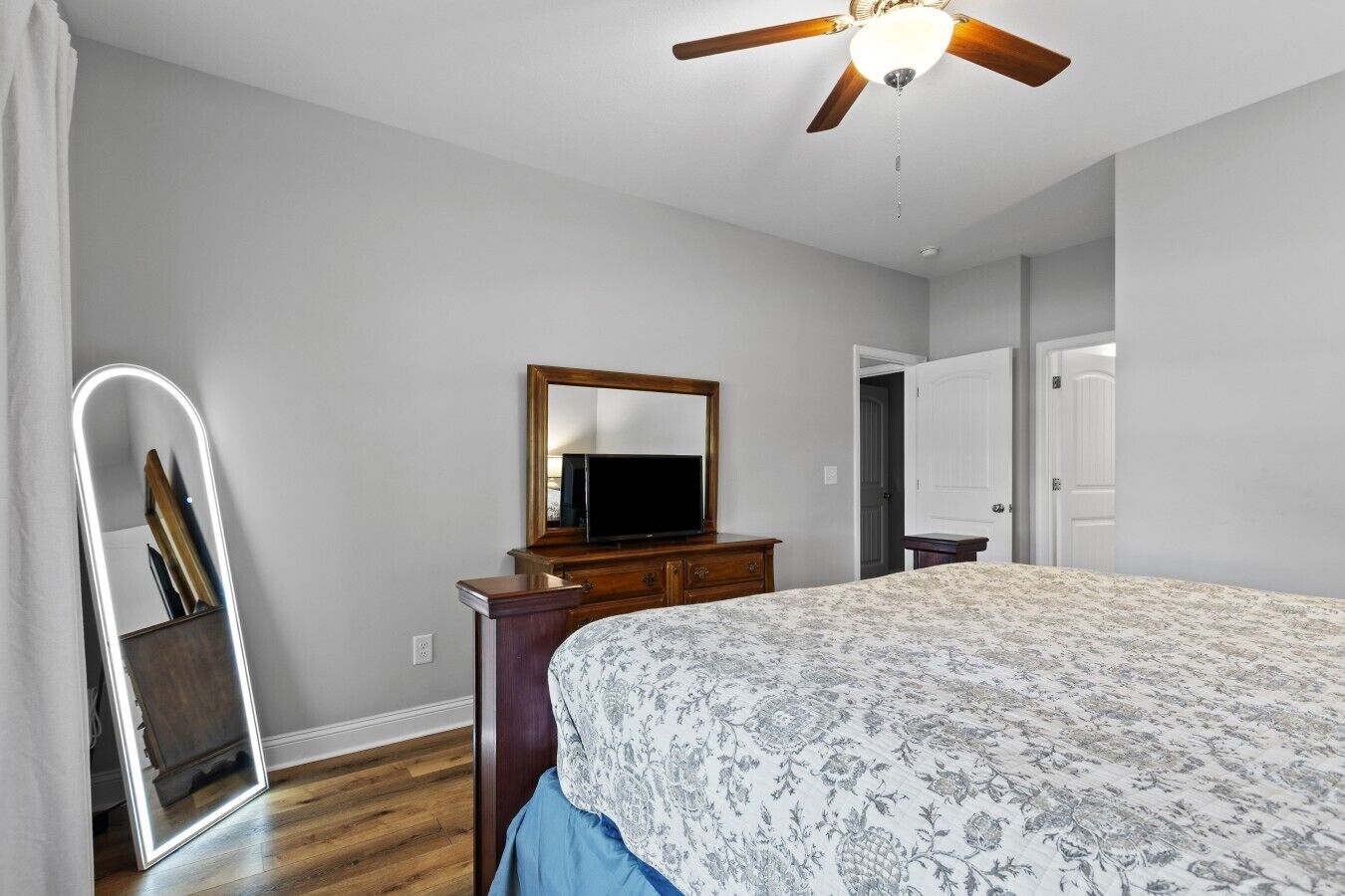 A bedroom with a bed, dresser, mirror, ceiling fan, and a door.