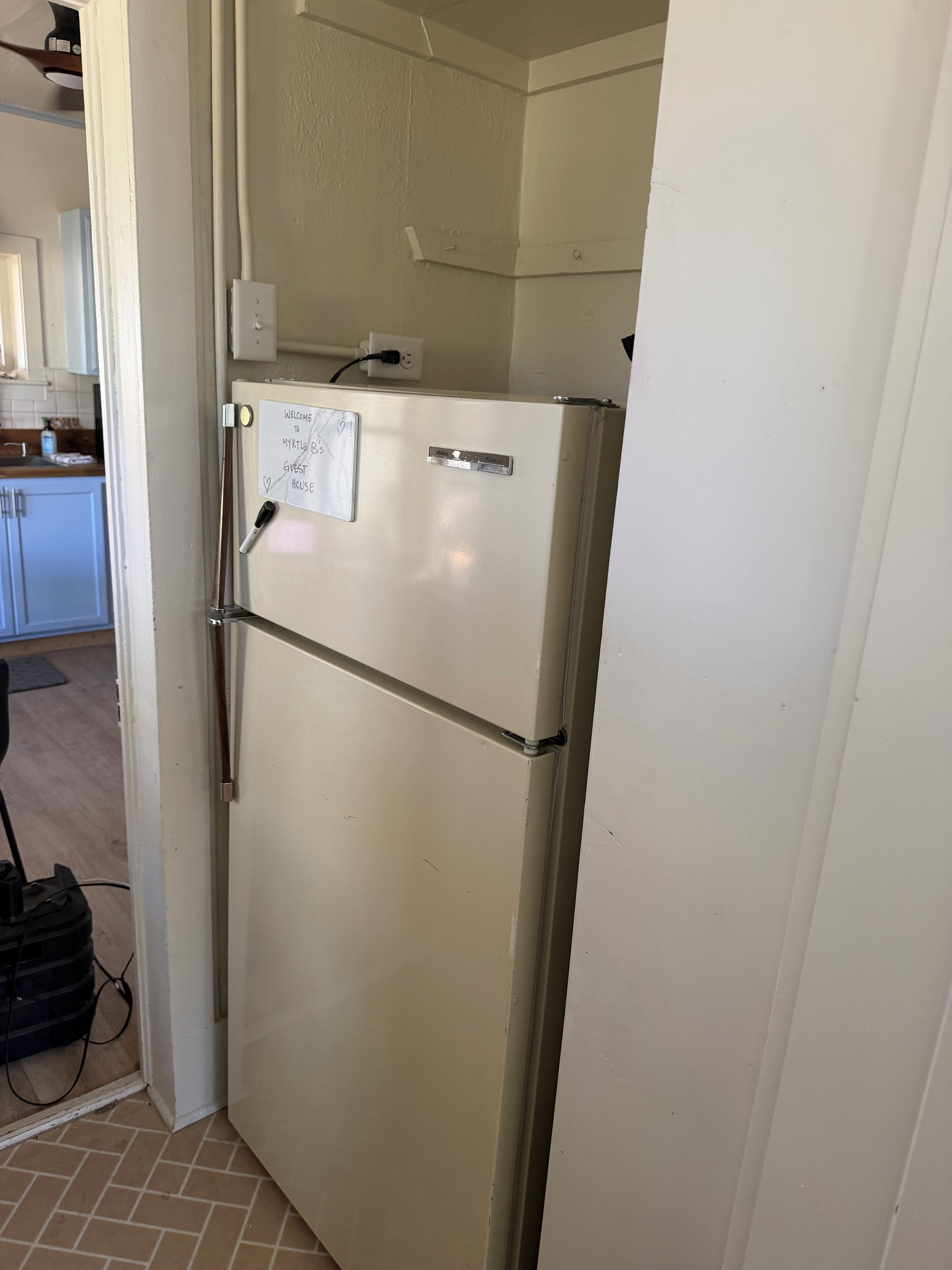 A kitchen with a white refrigerator, a wall-mounted electrical outlet, and a door with a handwritten note.