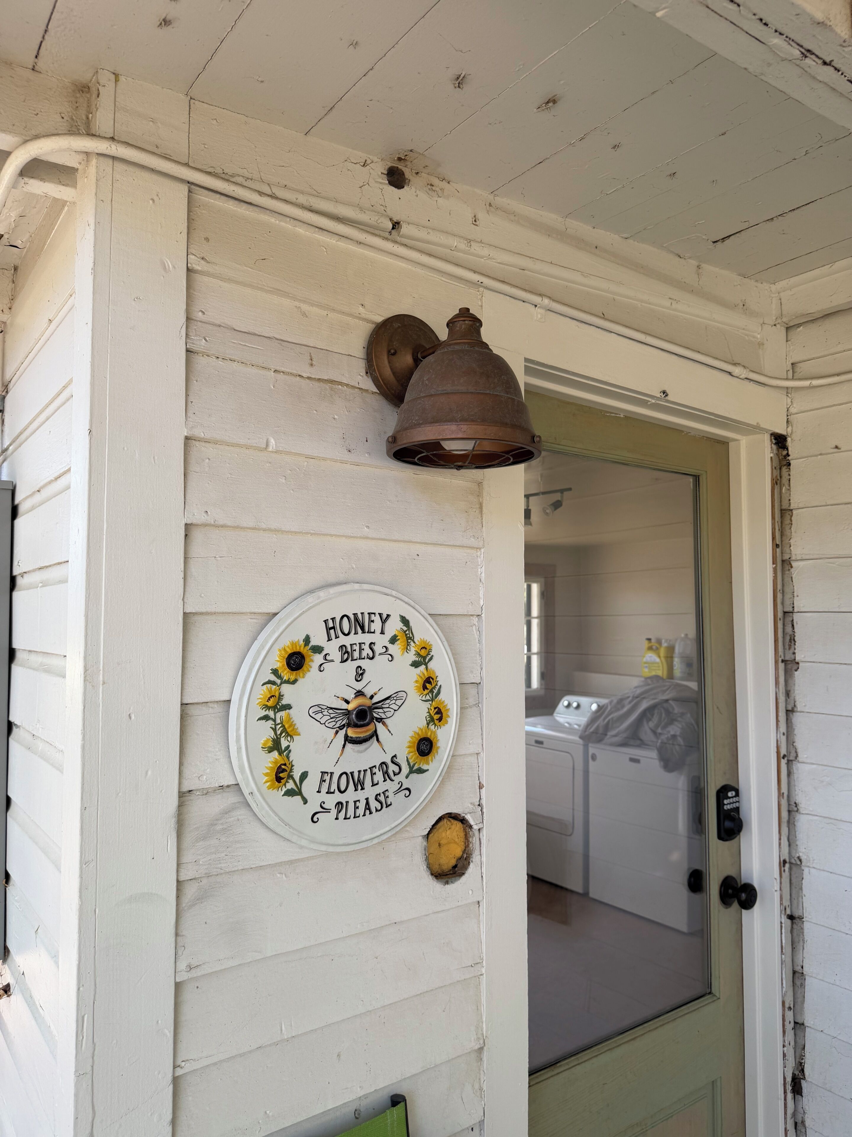 Un mur extérieur blanc, avec une porte usée et un panneau circulaire représentant des abeilles et des tournesols.