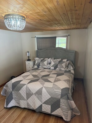 A bedroom with a wooden ceiling, a bed with a geometric patterned bedspread, a bedside table with a lamp, and a window with a view of greenery. - Wonderful 2-bedroom farmhouse in Salida.  Located @  Historic Hutchinson Ranch (Salida)