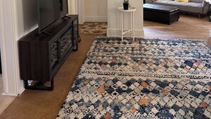 A living room with a flat-screen TV, a dark wooden entertainment center, a patterned rug, a white side table with a plant, and a grey sofa with a yellow pillow.