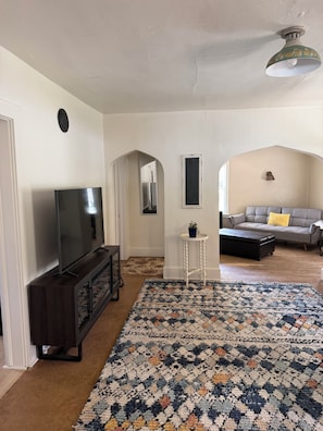 Sala de estar con televisor de pantalla plana, mueble de madera oscura para centro de entretenimiento, alfombra estampada, mesita auxiliar blanca con una planta y sofá gris con almohada amarilla.