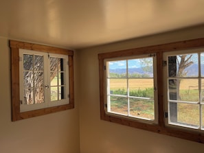 Una habitación con dos ventanas, una con vistas a un campo y montañas, y la otra con vistas a árboles.